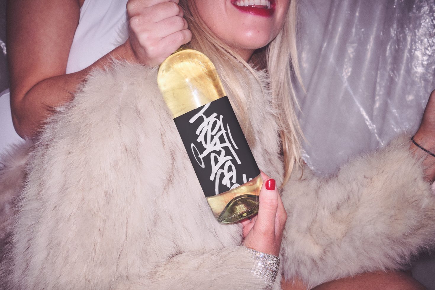 Happy woman holding a gold-labeled bottle mockup with stylish font, wrapped in a faux fur coat, showcasing product presentation.